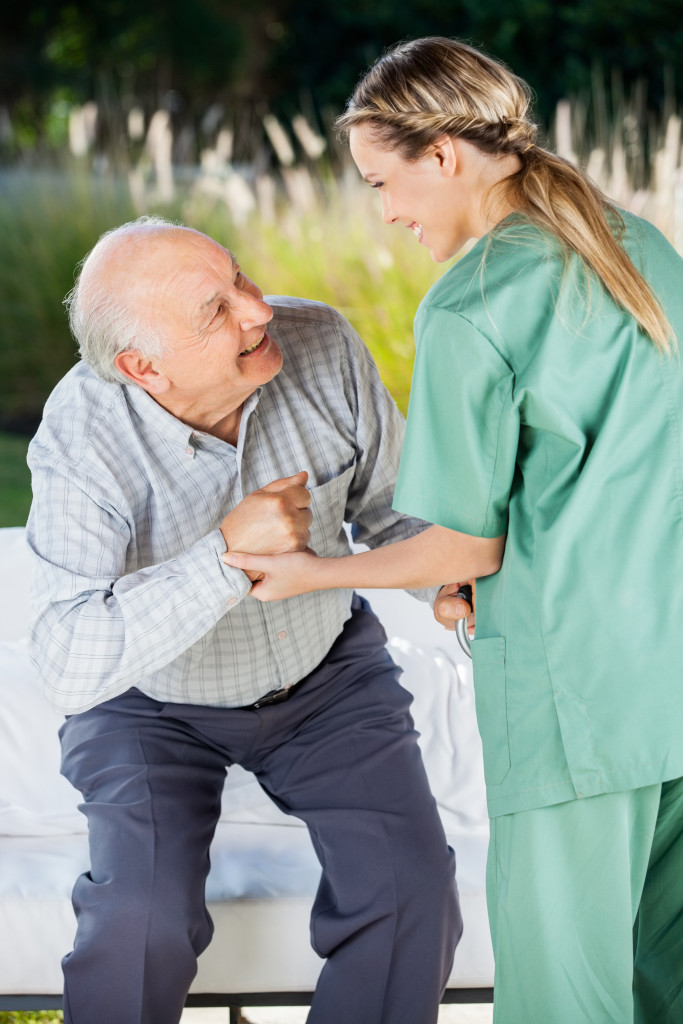 Female Nurse Helping Senior Man To Sit On Couch